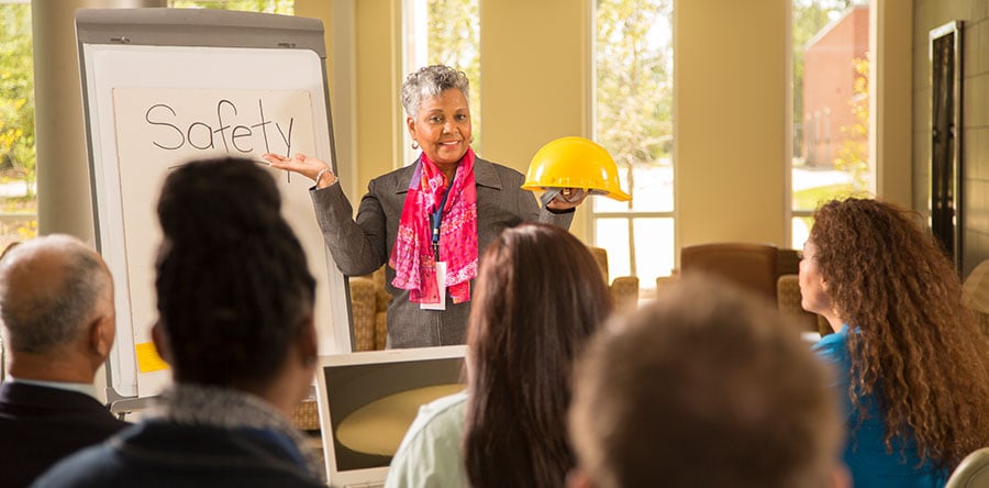 Instructor standing at front of classroom facing students holding yellow safety hat in left hand and right pointing to white board disussing safety.