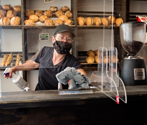 Image of a worker wearing a face mask standing behind a counter in a coffee shop with spray cleaner and cloth in hands wiping protective safety shield screen 