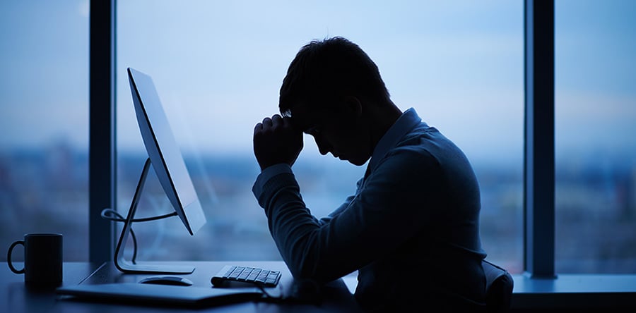 Man resting forehead on clasped hands and appearing stressed while sitting in front of his laptop