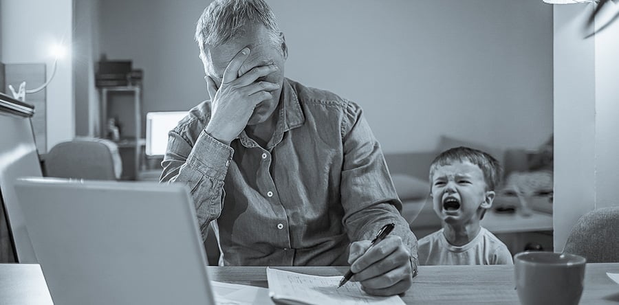 Black and white image of a man sitting at desk in front of computer with one hand covering face and next hand holding pen on a writing pad and crying child in background