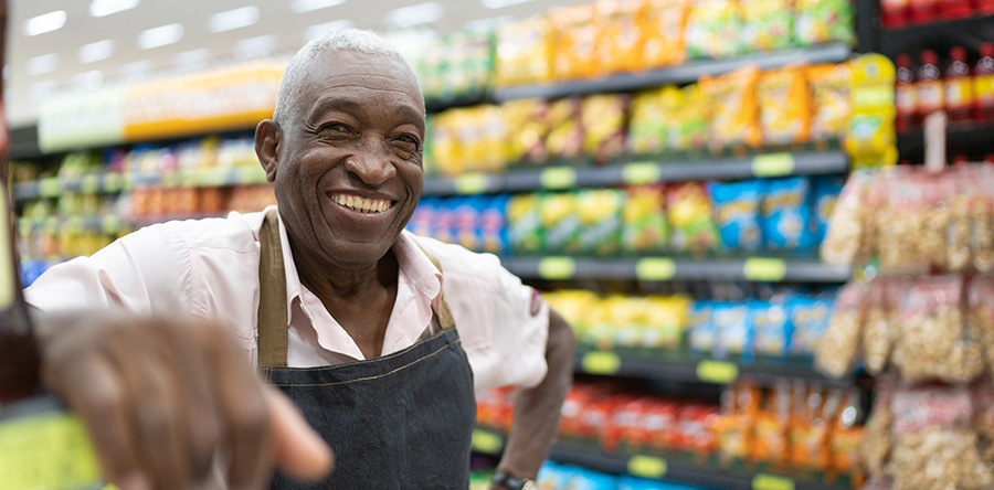 Grocery clerk wearing apron, standing in front of shelves, smiling happily at camera.