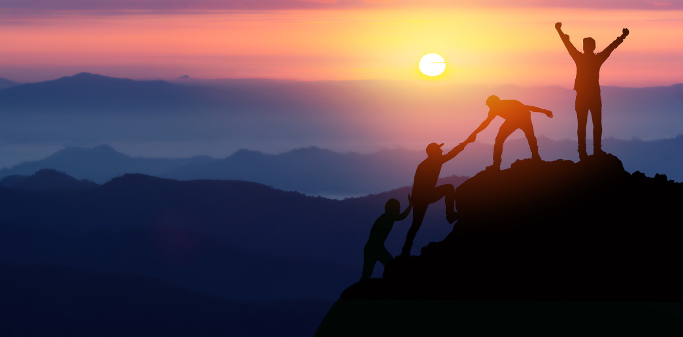 Image of individuals helping each other climb up a mountain during a sunrise.