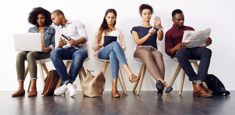 Image of people  sitting on chairs in a line looking at a book, phone, tablet, newspaper and laptop