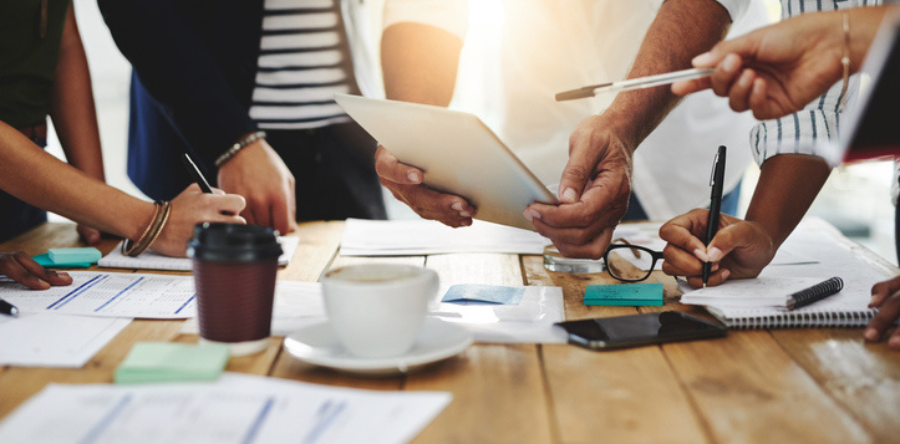 A group of office workers gathered around a wooden table looking at a paper document, discussing and writing notes with white paper, black pens, black cell phone, black glasses, white coffee mug, green and blue post it notes scattered around.  