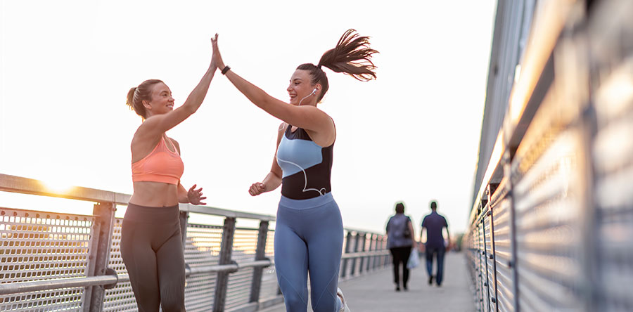Image of two woman wearing leggings, tank tops, hair in pony tail running on a pathway high fiving each other with one hand up in air 
