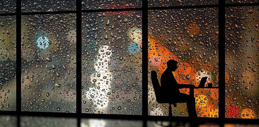 Image of a silhouette person sitting at workstation by the window in dark, rain drops on window with white, orange and blue light reflections off window.