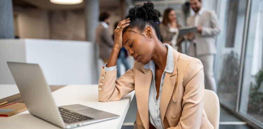 Image of woman feeling stressed and overwhelmed by work, holding her head with a hand, sitting at her office desk